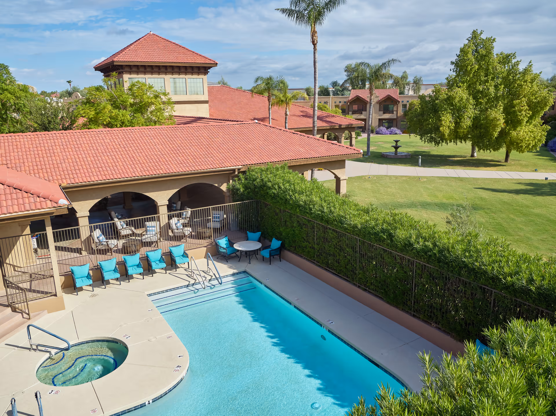 Outdoor swimming pool area with a hot tub, surrounded by a patio with blue cushioned chairs and tables. The pool area is adjacent to a building with a red tiled roof and arched openings. In the background, there is a large green lawn with trees, a fountain, and additional buildings with similar red tiled roofs under a partly cloudy sky.