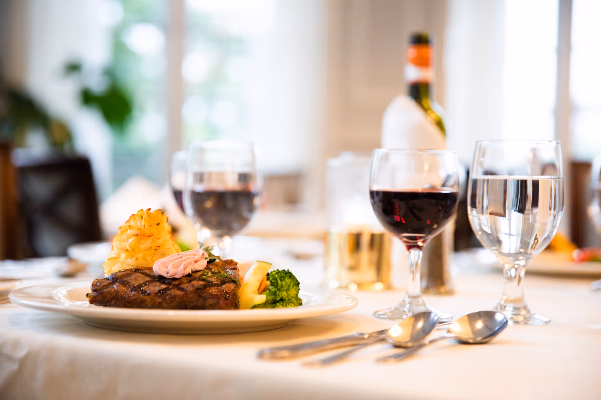 A plated meal featuring a grilled steak topped with a dollop of herb butter, a serving of mashed potatoes, and steamed broccoli on a white tablecloth. There are two glasses of red wine, a glass of water, and a bottle of wine in the background, with a softly lit dining room setting.