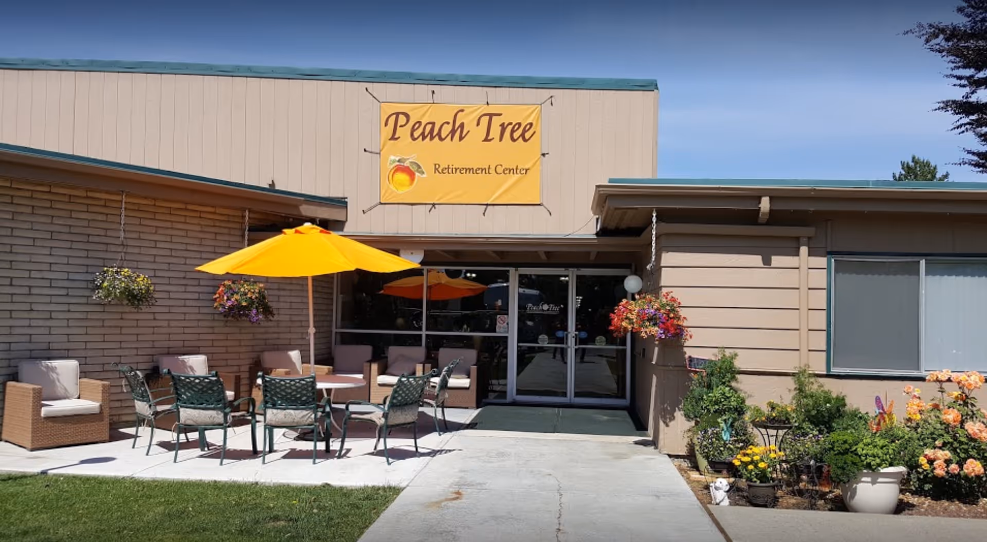 Exterior view of Peach Tree Retirement Center showing the entrance with glass double doors. There is a yellow sign above the entrance with the facility name and a peach graphic. Outside the entrance, there is a patio area with outdoor seating including cushioned chairs, metal chairs, tables, and yellow umbrellas. Hanging flower baskets and a garden with various flowers and plants are visible on the right side.