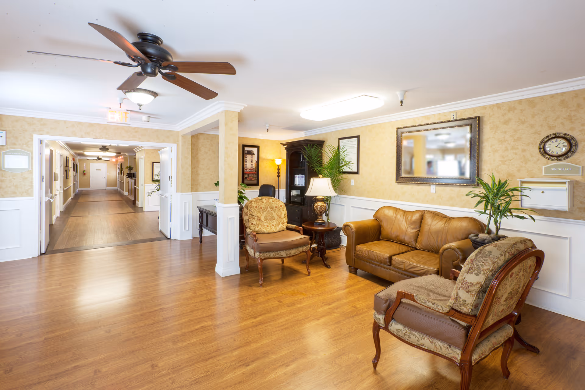 A well-lit common area in a senior living facility featuring a brown leather sofa, two upholstered armchairs, a wooden side table with a lamp, and decorative plants. The room has wood flooring, beige patterned wallpaper with white wainscoting, a ceiling fan, and a hallway leading to other rooms.