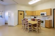 Bright institutional kitchen with a central dining table and chairs, white appliances, and light wood cabinetry.