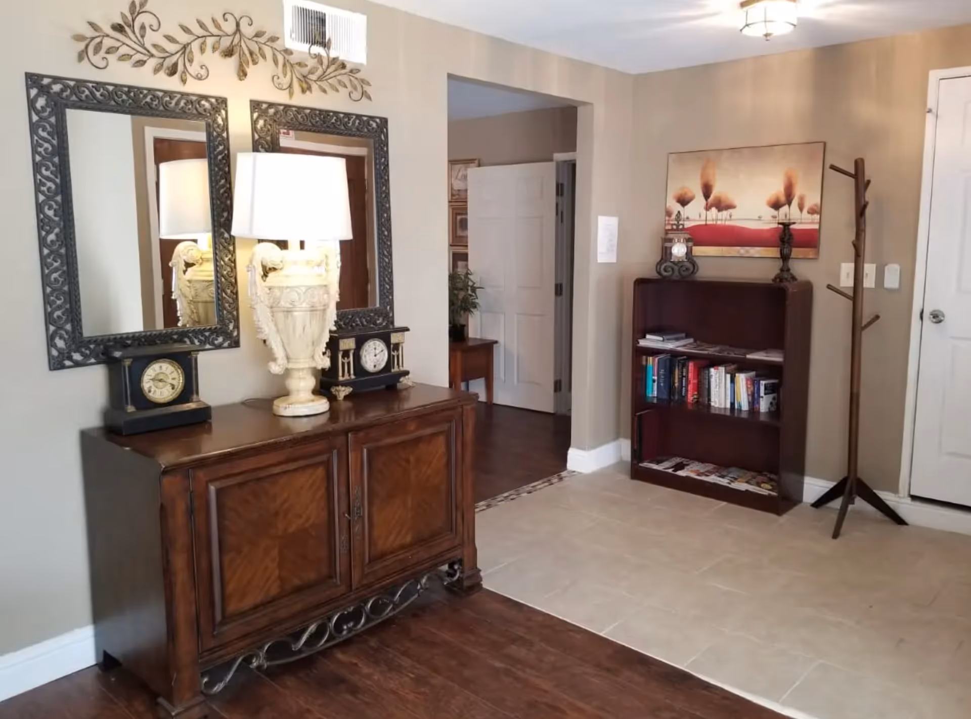 Interior view of a room with a wooden cabinet topped with two decorative clocks and a large ornate lamp, two framed mirrors on the wall above the cabinet, a bookshelf filled with books and magazines, a coat rack, and a painting of trees on the wall. The floor transitions from wood to tile near a doorway leading to another room.