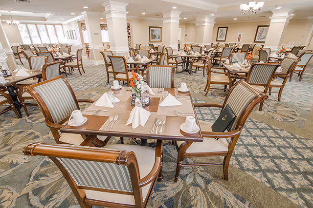 A spacious dining room with multiple wooden tables and chairs arranged neatly. Each table is set with white napkins folded in a triangular shape, white cups, saucers, and silverware. The room has a patterned carpet and is well-lit with natural light coming through large windows on the left side. The walls are decorated with framed artwork and the ceiling has recessed lighting and chandeliers.