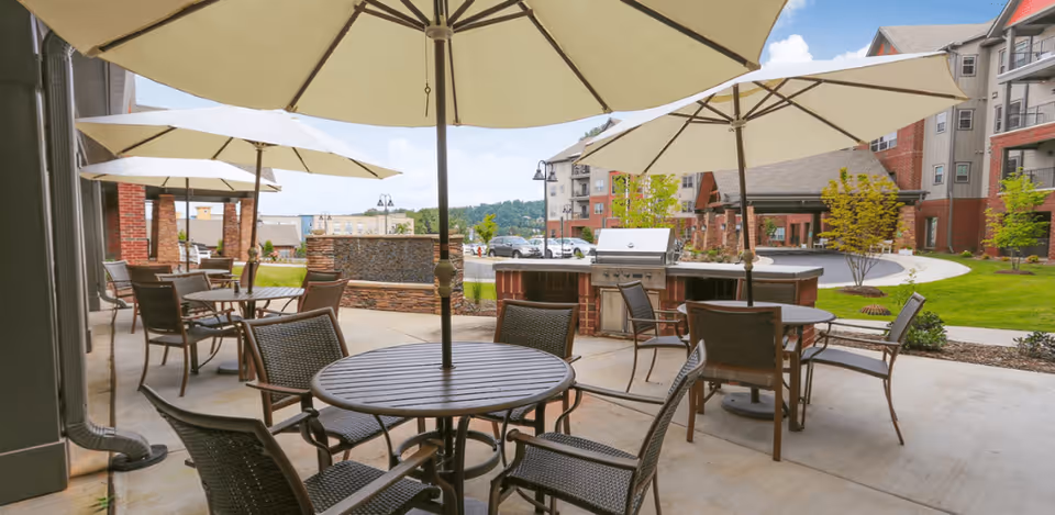Outdoor patio area with round tables and chairs under large white umbrellas. There is a built-in grill and a water feature with a stone facade. In the background, there are multi-story residential buildings and a well-maintained lawn with trees and shrubs.