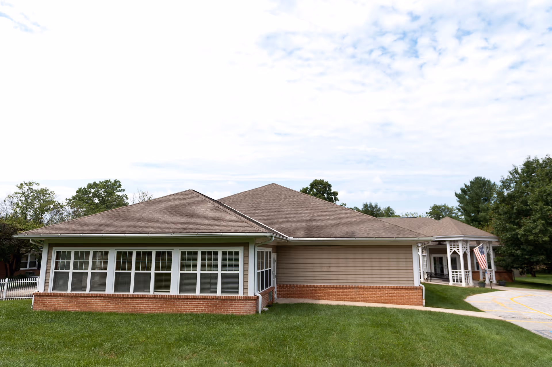 Exterior view of a single-story nursing and rehabilitation center building with a brown shingled roof, beige siding, and a brick foundation. The building has multiple windows with white frames and a small covered porch with white railings and an American flag. The surrounding area includes green grass and trees under a partly cloudy sky.