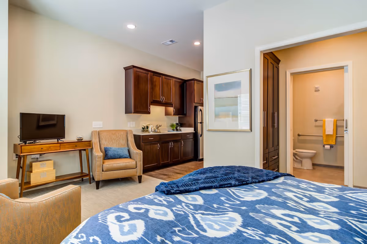 A senior living facility room featuring a bed with a blue and white patterned blanket in the foreground, a beige armchair with a blue cushion, a wooden console table with a small TV, and a kitchenette with dark wood cabinets and a refrigerator. In the background, there is a bathroom with a toilet and a yellow towel hanging on the wall.