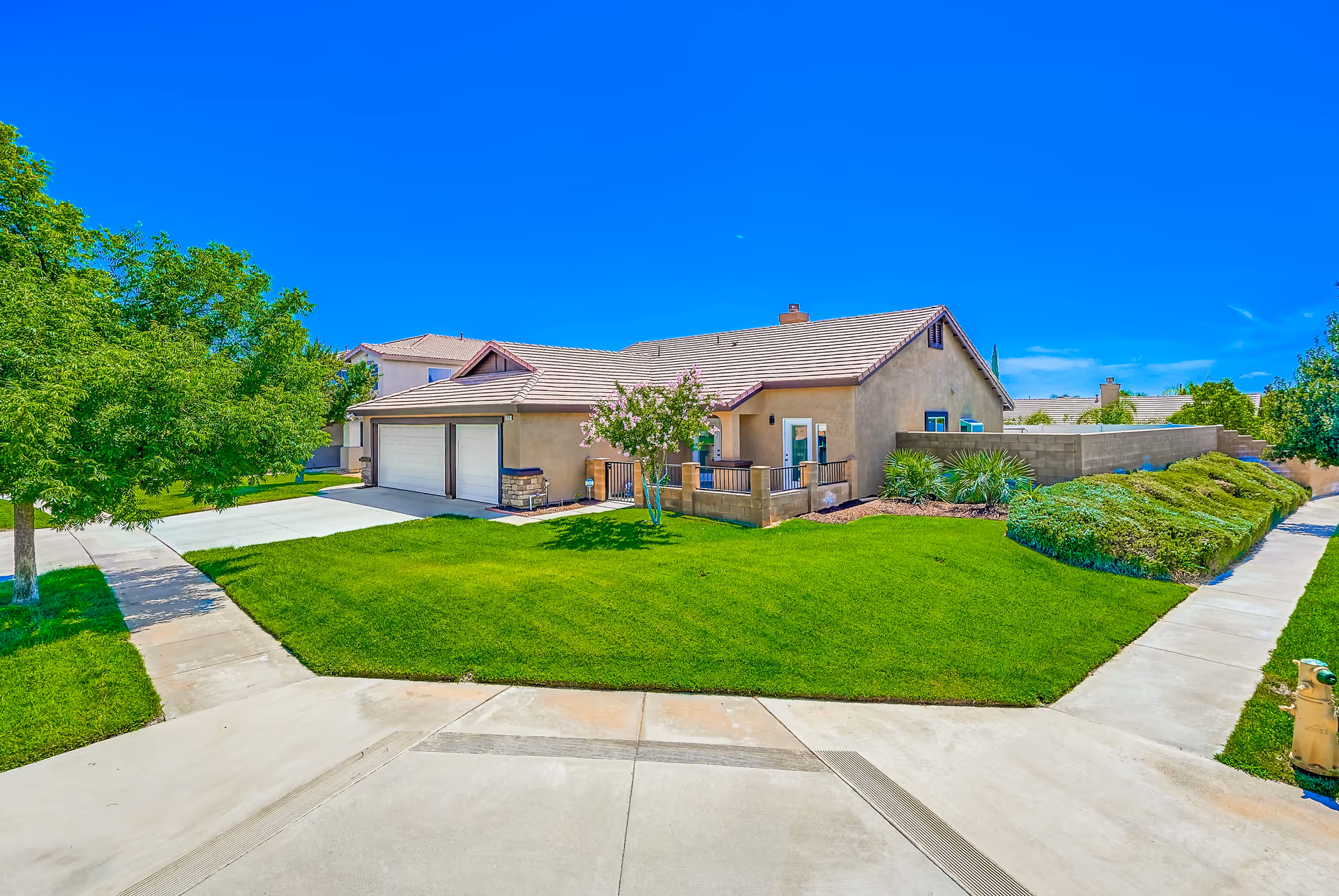 Exterior view of a single-story residential building with a beige stucco finish and a tiled roof, surrounded by a well-maintained green lawn, trees, and shrubs under a clear blue sky. There is a concrete driveway leading to a two-car garage and sidewalks bordering the property.
