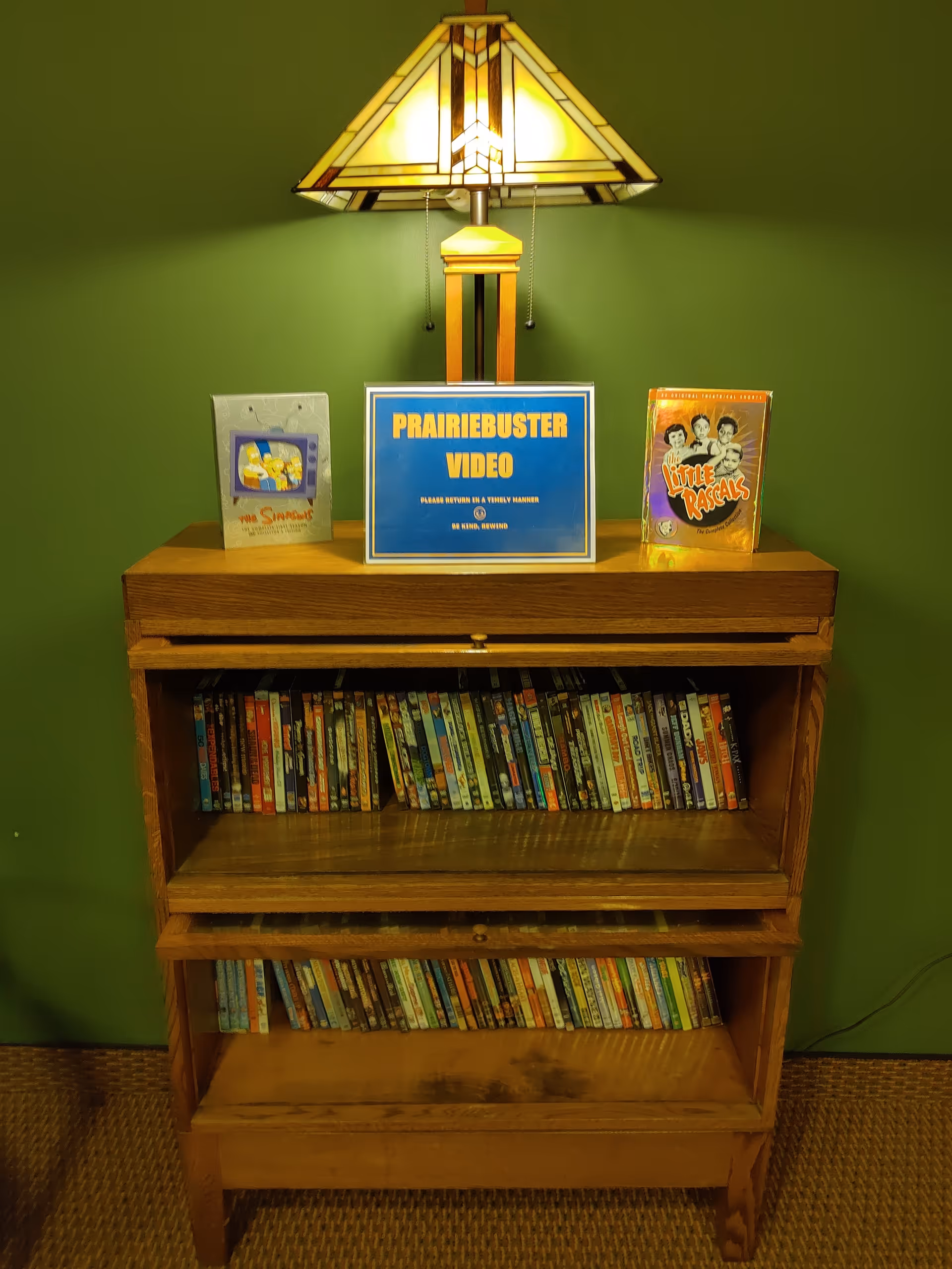 A wooden media cabinet against a green wall with two shelves filled with DVDs. On top of the cabinet is a stained glass table lamp and three items: a DVD case of The Simpsons, a sign that reads 'PRAIRIEBUSTER VIDEO PLEASE RETURN IN A TIMELY MANNER BE KIND, REWIND', and a DVD case of Little Rascals.