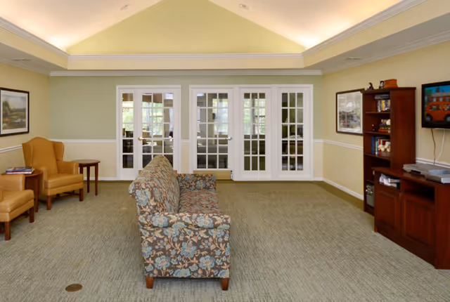 A well-lit living room with a floral patterned sofa in the center, two mustard yellow armchairs with a small wooden side table on the left, and a wooden entertainment unit with a TV and shelves on the right. The room has light green walls with white trim, a high vaulted ceiling with recessed lighting, and three white French doors at the back leading to another room.