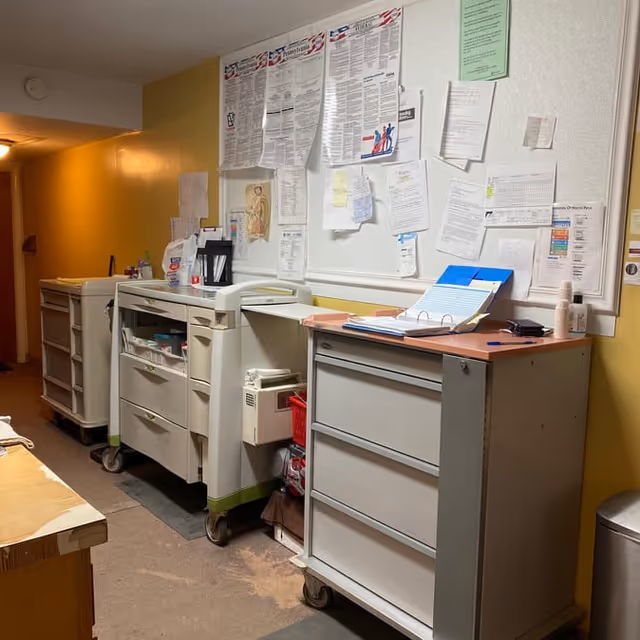 A hallway area in a senior care facility with medical carts and filing cabinets on wheels. The wall behind the carts is covered with various posted notices, papers, and schedules. The walls are painted yellow and white, and the floor appears to be a mix of carpet and linoleum.