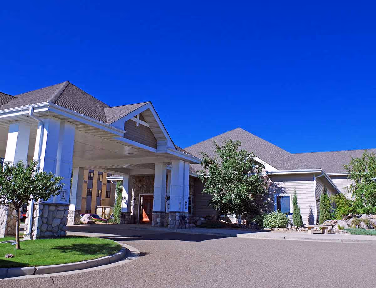 Front entrance of a single-story senior living facility with a covered porte-cochere, stone pillars, and landscaping under a clear blue sky.
