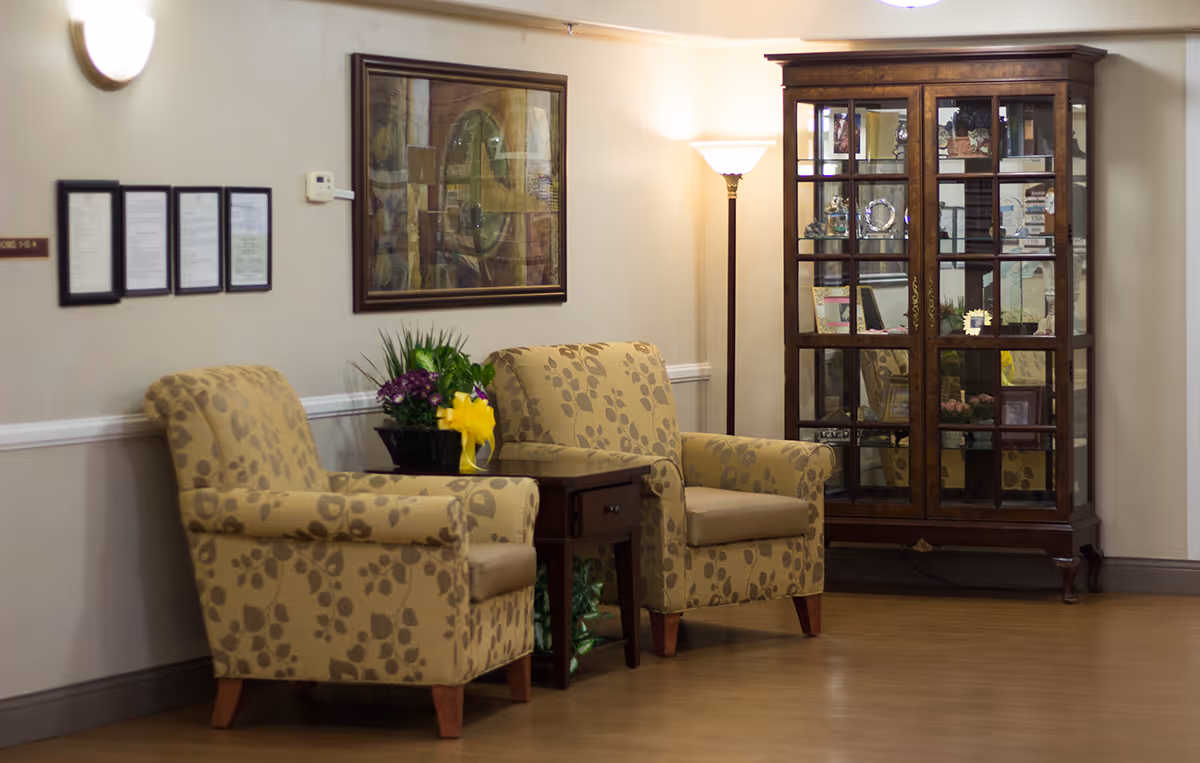 A cozy seating area with two beige armchairs featuring a leaf pattern, separated by a small wooden side table with a flower arrangement. Behind the chairs is a framed artwork on the wall and a tall floor lamp providing warm light. To the right, there is a wooden glass-front cabinet displaying various decorative items and awards.