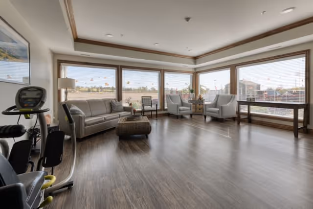 Sunlit common lounge with a sofa, armchairs, exercise equipment and large windows overlooking the outdoors.