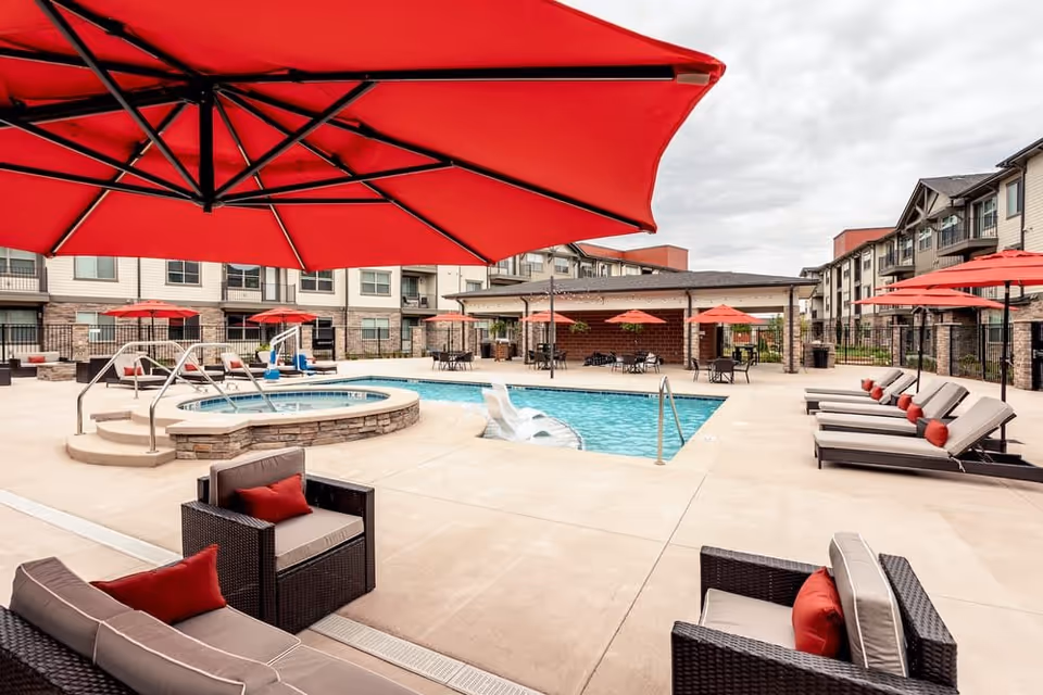 Outdoor pool area at Eagle's Peak facility featuring a swimming pool, a hot tub, several lounge chairs with red cushions, multiple large red umbrellas providing shade, and a covered seating area in the background. The surrounding buildings are multi-story with balconies.