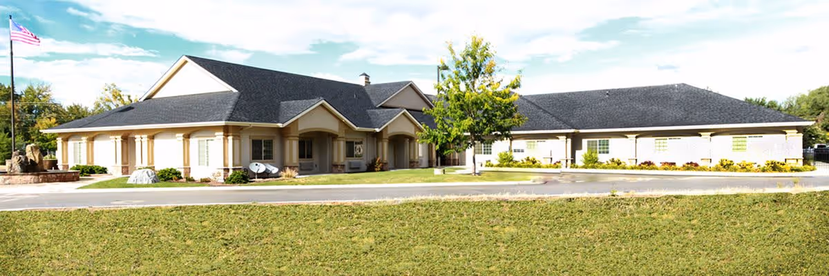 Exterior view of a single-story building with beige walls and a dark gray roof, surrounded by a grassy area and a paved driveway. There is an American flag on a flagpole to the left and some landscaping with bushes and a tree in front of the building.
