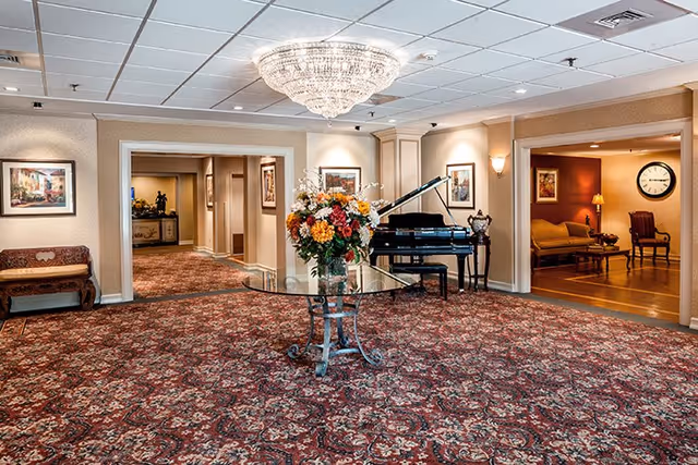 Spacious carpeted interior lounge with a crystal chandelier, a glass table topped with a floral arrangement, and a grand piano near seating alcoves.