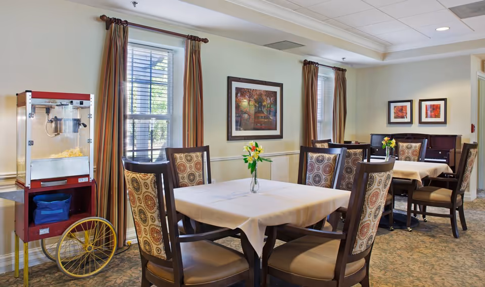 A dining room with two tables covered in white tablecloths, each surrounded by four wooden chairs with patterned cushions. There are vases with yellow flowers on each table. The room has two windows with striped curtains, framed artwork on the walls, and a popcorn machine on a cart with yellow wheels near one window.