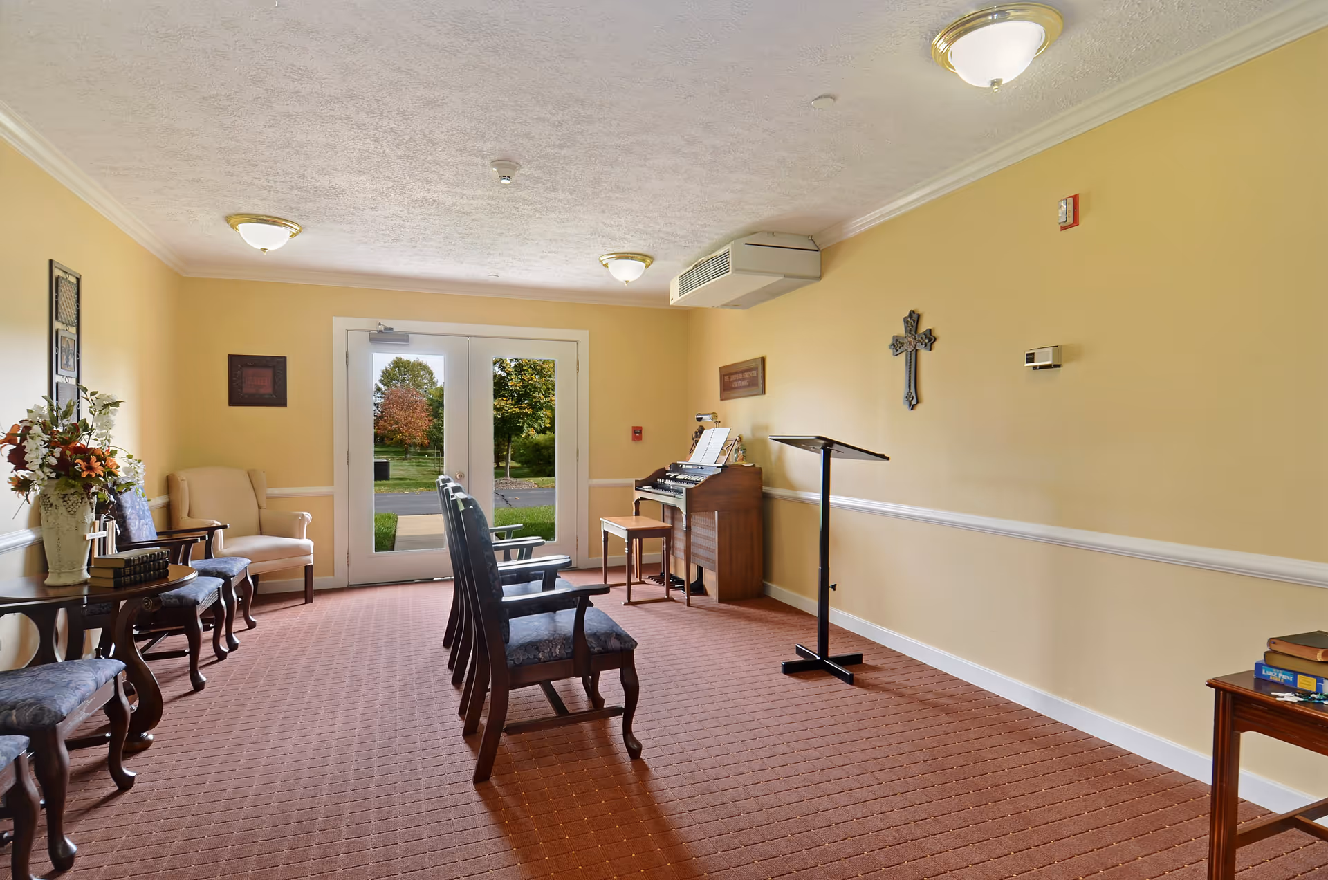 A small room with beige walls and carpeted floor featuring several wooden chairs with blue cushions arranged in two rows facing a wooden organ and a music stand. There is a double glass door at the far end showing a view of green trees outside. The room is decorated with a vase of flowers on a side table, framed pictures on the walls, and a cross hanging on the right wall.