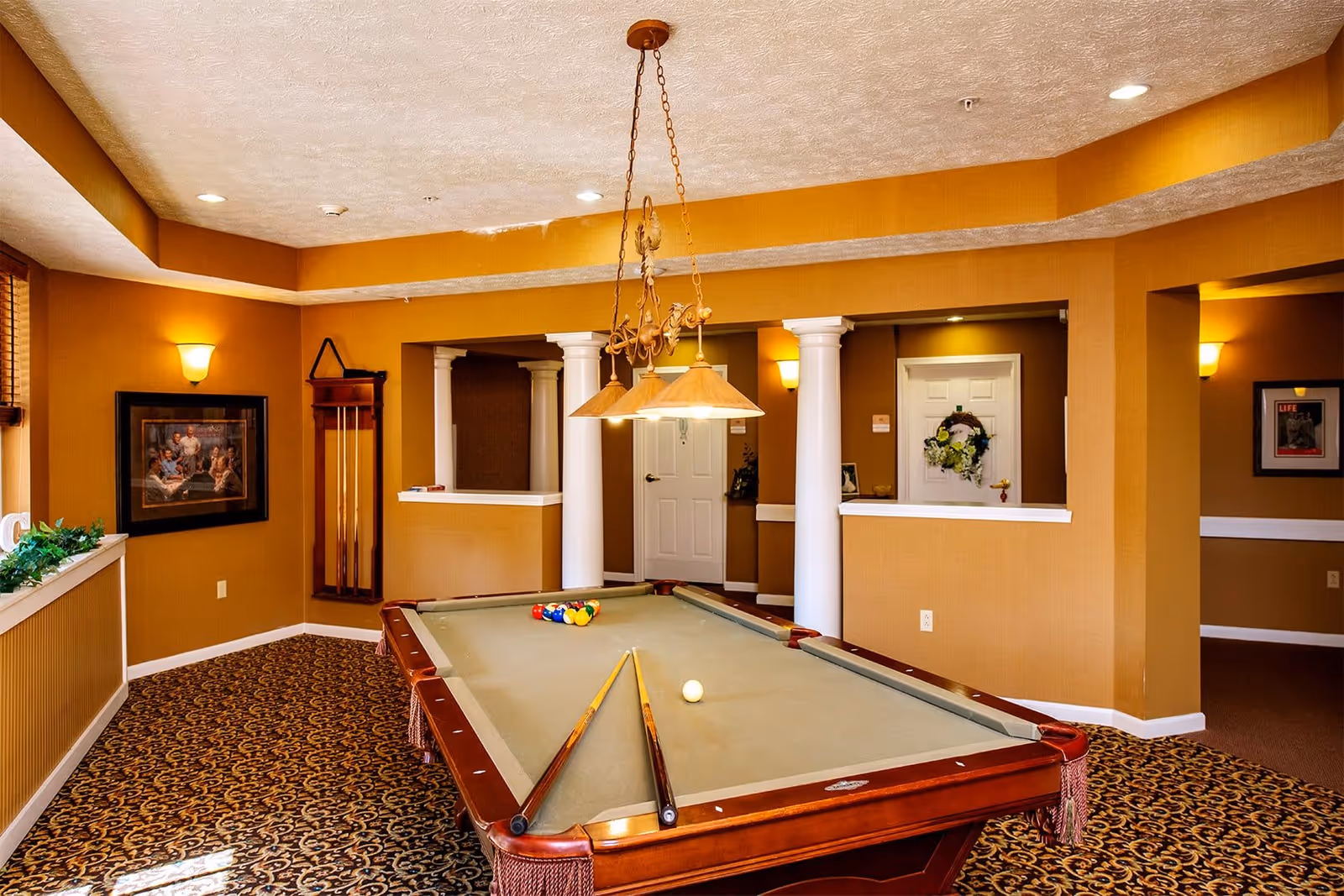 Interior room with a pool table in the center, two pool cues resting on the table, and a rack of pool cues mounted on the wall. The room has warm yellowish-brown walls, patterned carpet, white columns, framed pictures on the walls, and a hanging light fixture above the pool table.