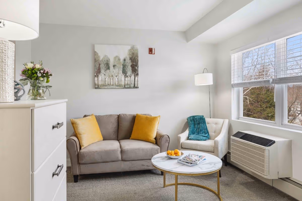A cozy living room with a gray loveseat adorned with two yellow pillows, a white armchair with a teal throw blanket, a round marble coffee table with a bowl of oranges and magazines, a white dresser with a lamp and a vase of flowers, a floor lamp, and a large window showing trees outside.
