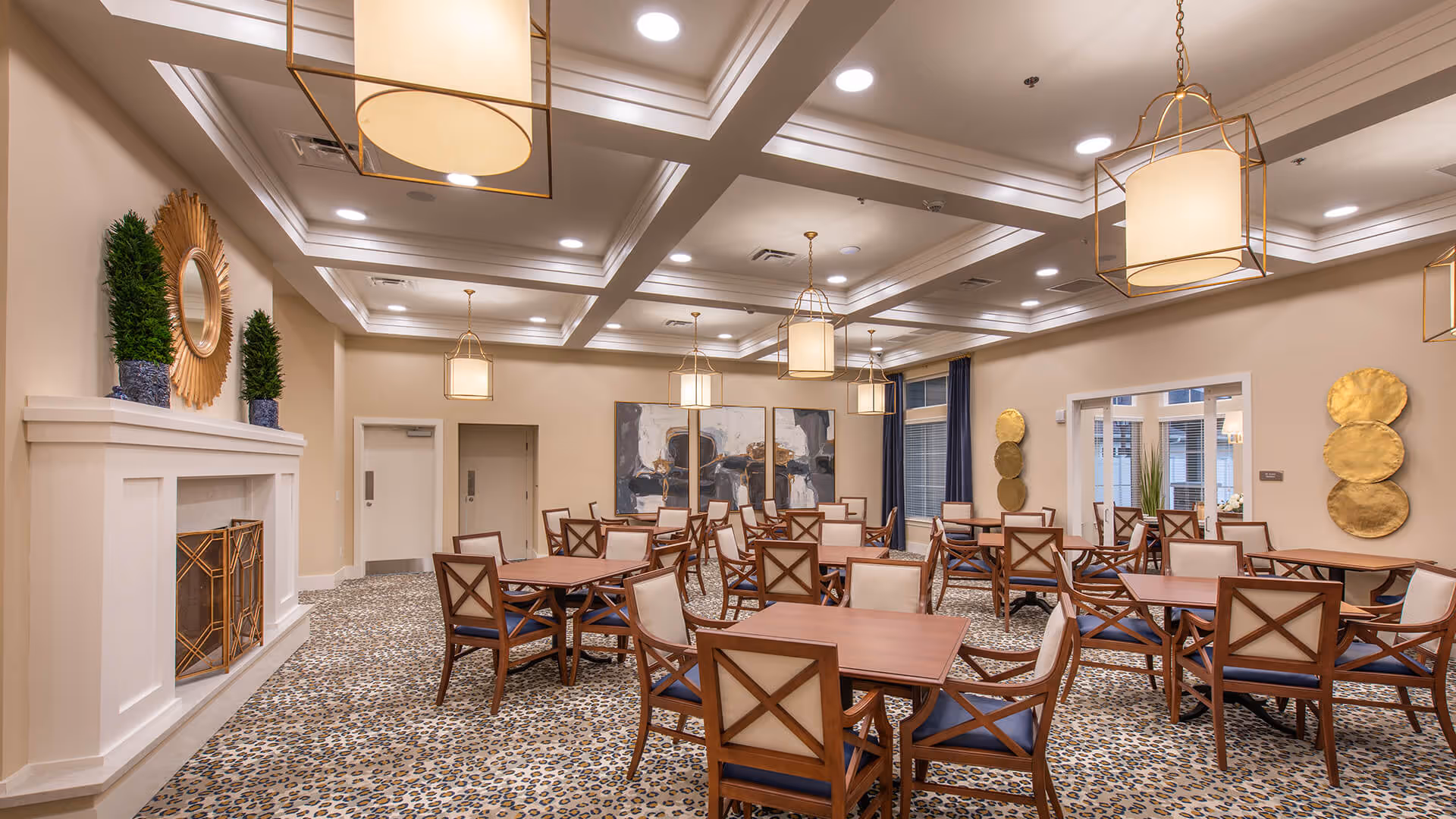 Spacious, well-lit communal dining room with multiple wooden tables and chairs, decorative pendant lights, a fireplace, and wall art.