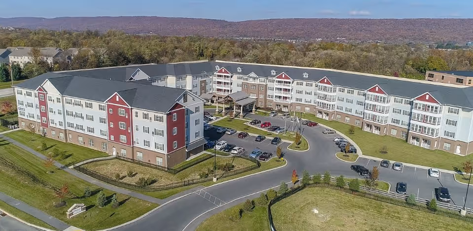 Aerial view of a large senior living facility named Harmony at West Shore, featuring a multi-story building with white and red exterior walls, surrounded by parking lots, green lawns, and trees with a backdrop of forested hills under a clear sky.