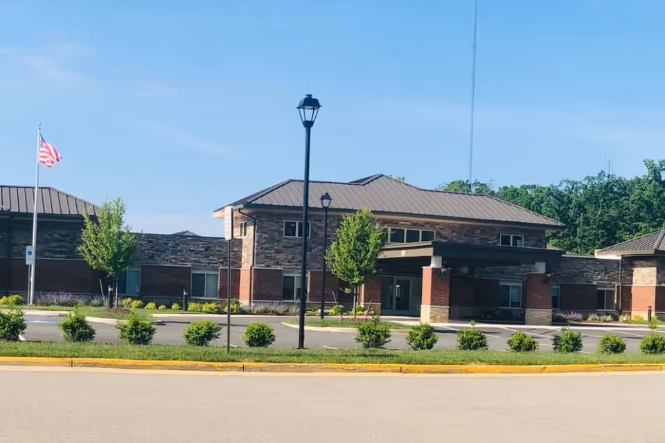 Front exterior of a brick-and-stone memory care building with a covered entrance, flagpole, and landscaped parking area.