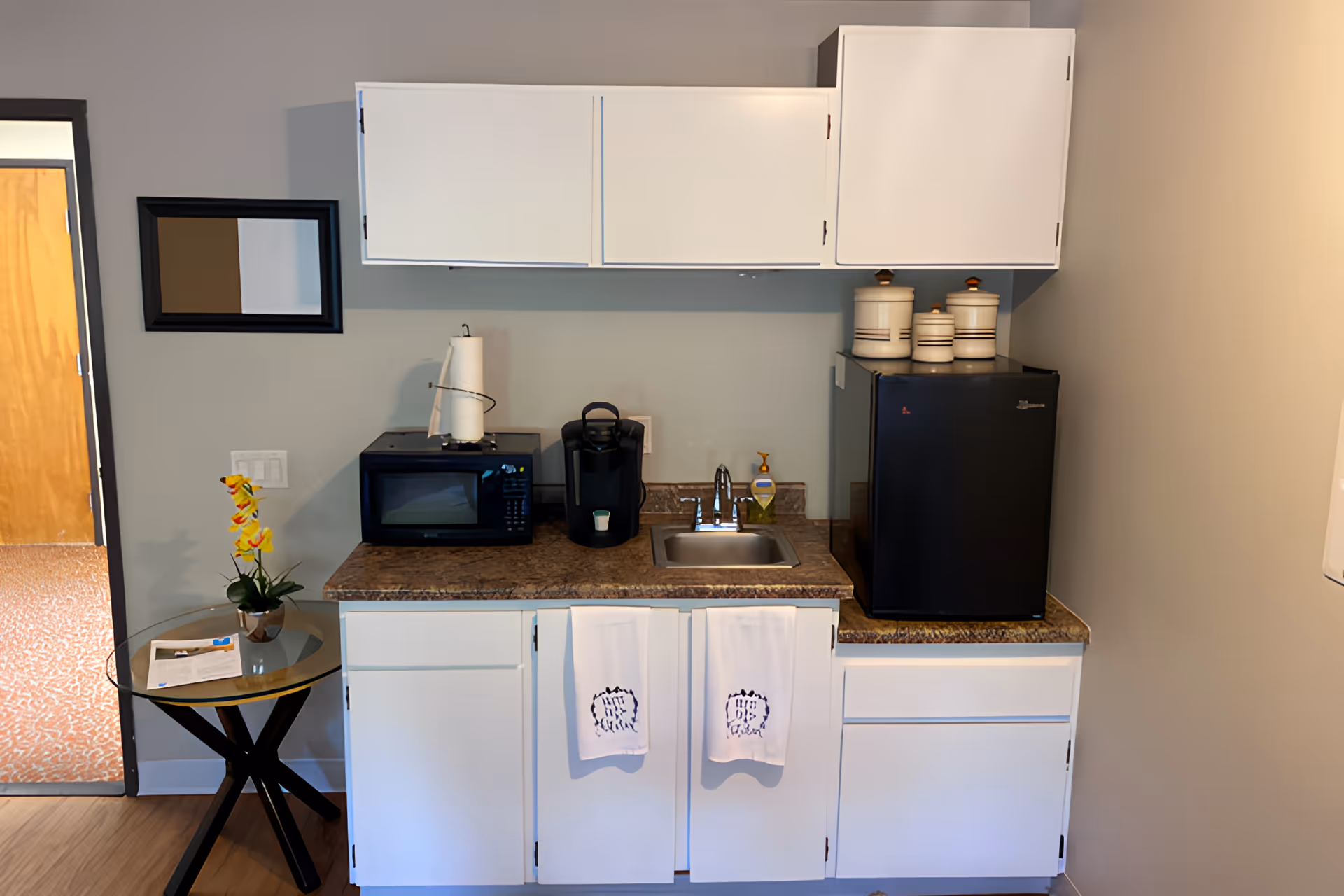 A small kitchenette area with white cabinets, a countertop with a microwave, coffee maker, small sink, and a black mini refrigerator. Two white towels hang on the cabinet doors below the sink. To the left, there is a small round glass table with a yellow flower in a pot and some papers on it. A rectangular mirror is mounted on the wall above the microwave.