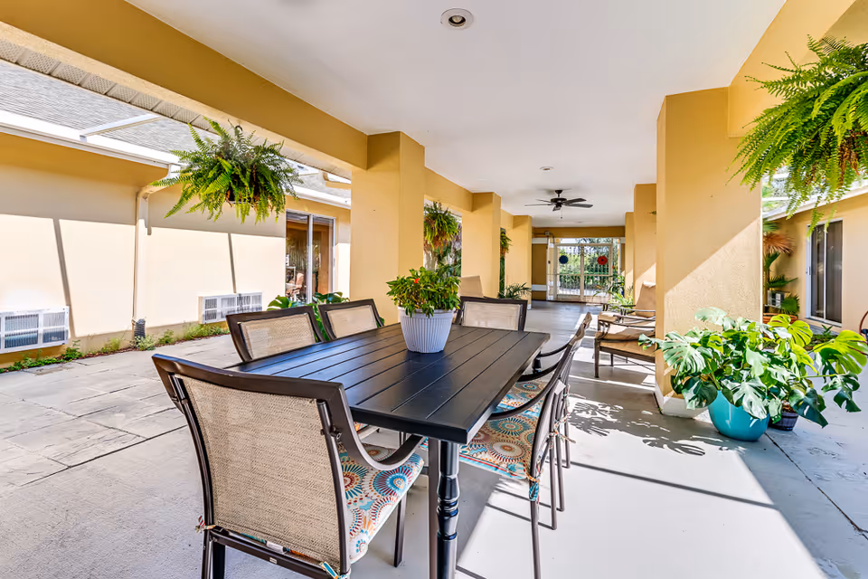 Covered outdoor patio area with a black rectangular table surrounded by six chairs with patterned cushions. Several green potted plants and hanging ferns decorate the space. The area is shaded by a yellow-beige structure with ceiling fans and opens to an outdoor garden area.
