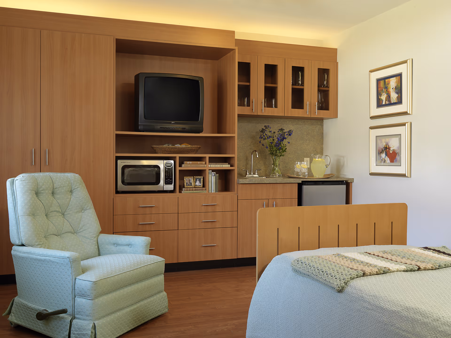 A cozy room with a light blue upholstered recliner chair, a wooden bed with a crocheted blanket, and a built-in wooden cabinet unit. The cabinet includes a small TV, microwave, shelves with books and picture frames, a sink with a vase of flowers, a pitcher of lemonade with glasses, and a mini refrigerator. Two framed paintings hang on the wall above the bed.