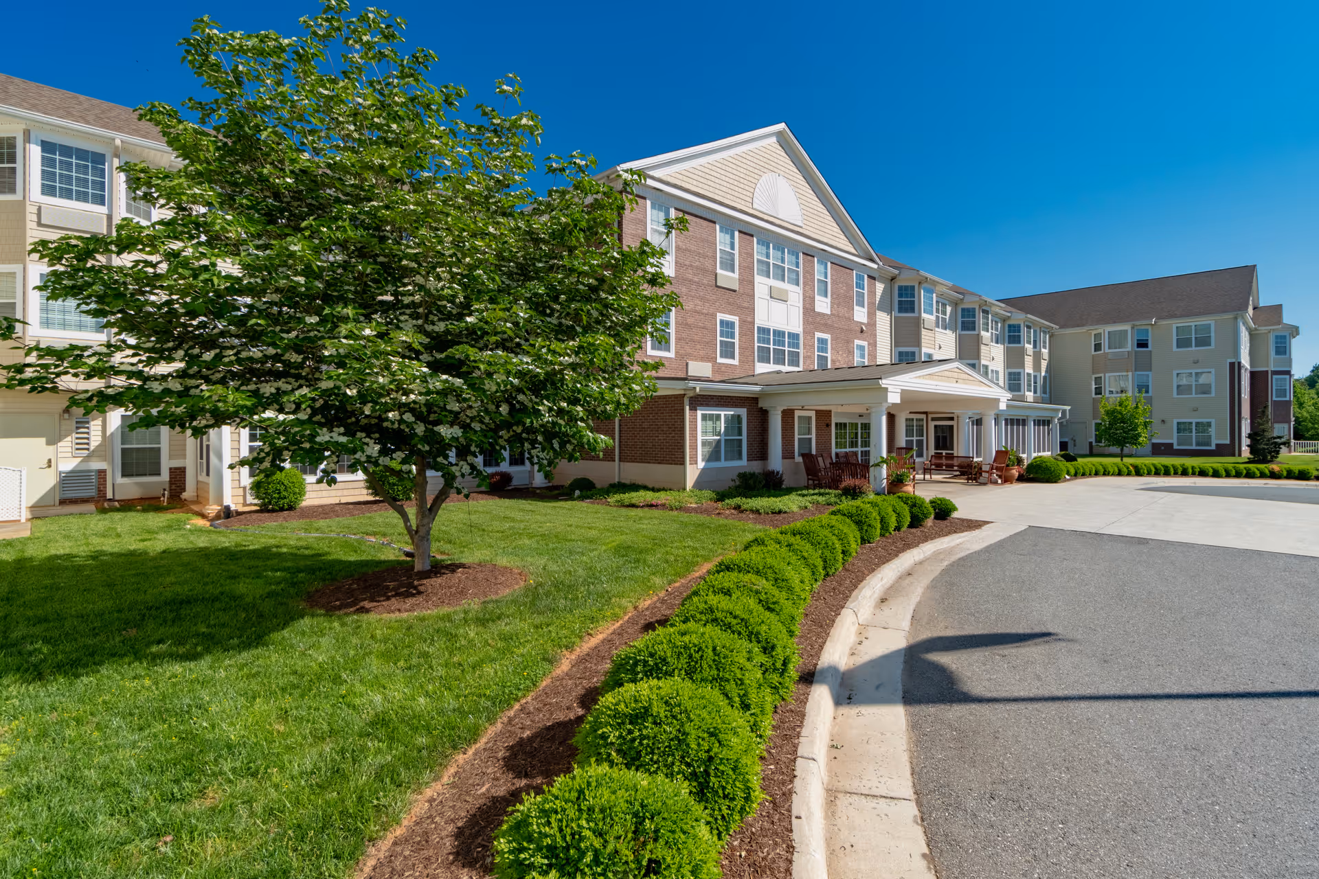 Front exterior of a multi-story senior living facility with a covered entrance, landscaped lawn, shrubs, and a tree.