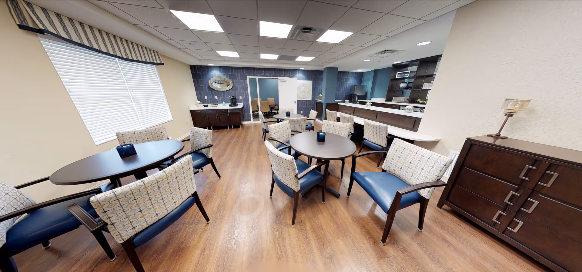 A bright and spacious common dining area in a senior living facility featuring multiple round tables with cushioned chairs and benches. The room has wood flooring, a large window with blinds, and a counter area with cabinets and shelves in the background. The walls are painted in neutral tones with a decorative blue accent wall.