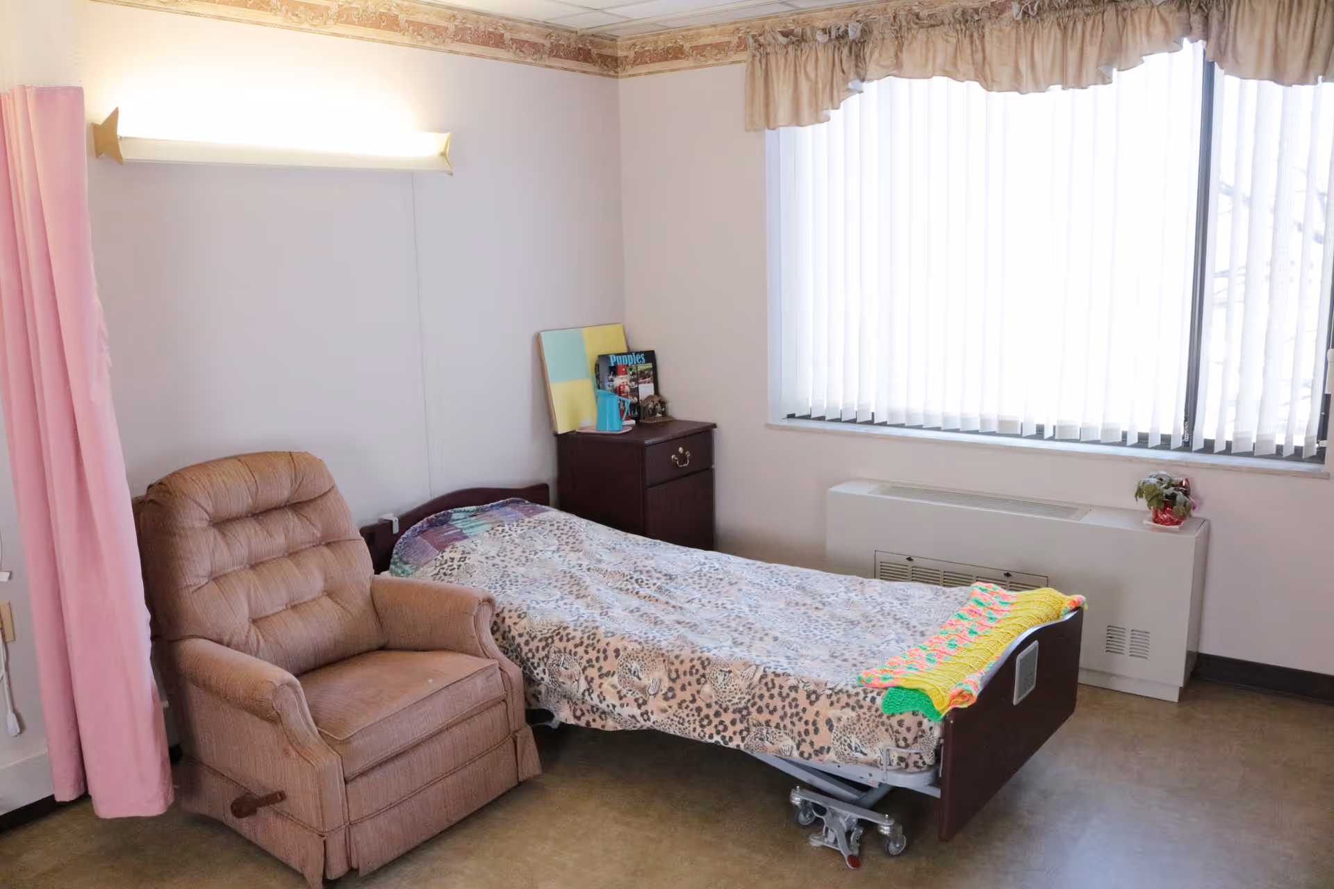 A simple bedroom in a senior living facility featuring a single bed with a patterned blanket and a colorful knitted throw at the foot. Next to the bed is a brown recliner chair and a small dark wooden nightstand with books and a small decorative item. The room has a large window with vertical blinds and a heating unit below it. The walls are light-colored with a floral border near the ceiling, and there is a pink curtain partially visible on the left side.