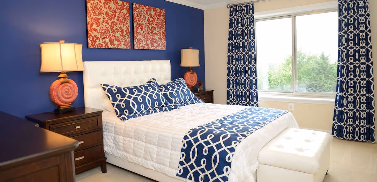 A bedroom with a white upholstered bed featuring blue and white patterned pillows and a matching runner. There are two wooden nightstands with coral-colored lamps on either side of the bed. The wall behind the bed is painted blue and decorated with two red and white patterned artworks. Blue and white patterned curtains frame a large window letting in natural light. A white tufted bench is placed at the foot of the bed.