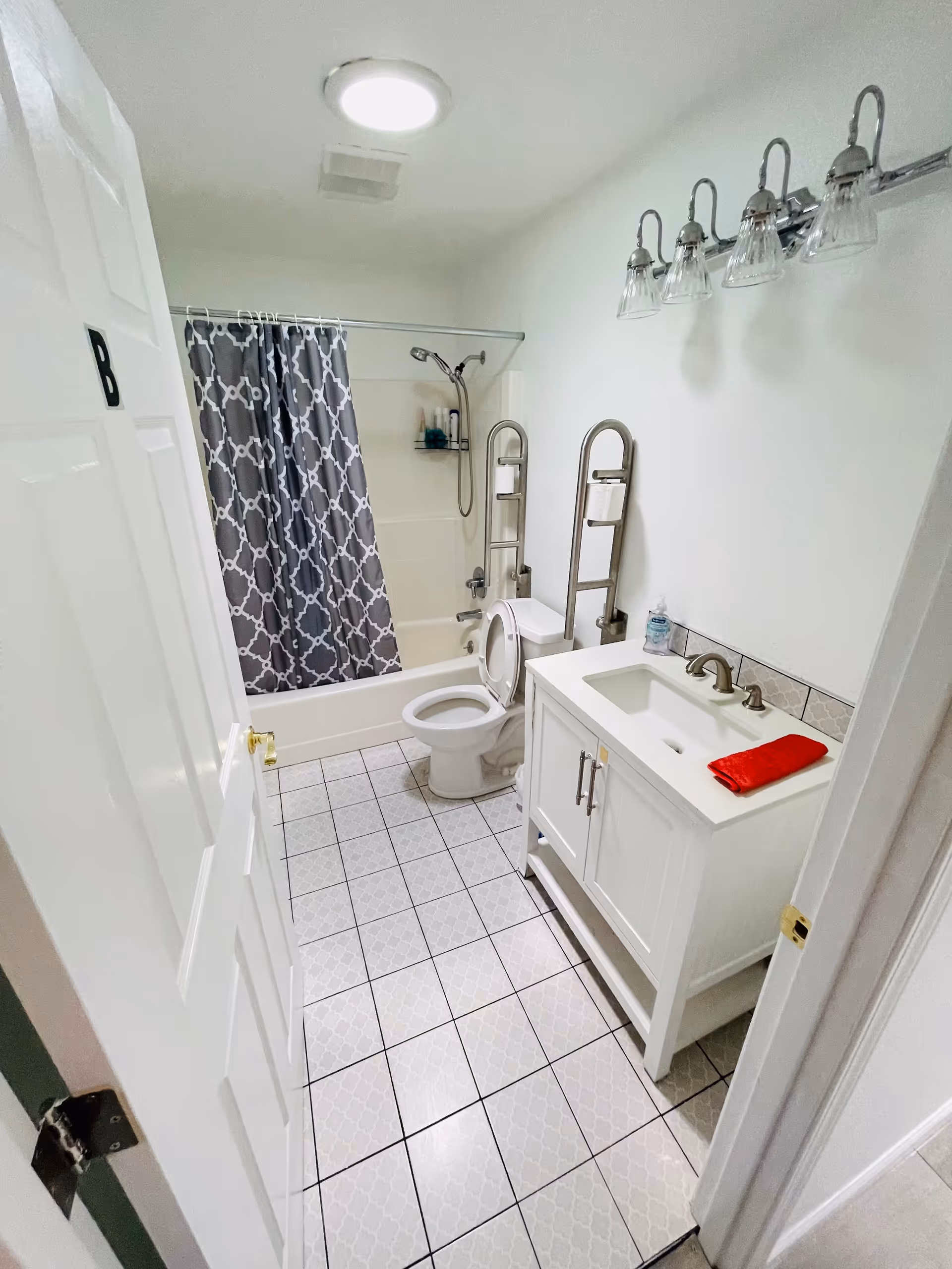 A clean and bright bathroom with white tiled floor and walls. There is a white vanity with a sink and a red towel on it, a toilet with safety grab bars on both sides, and a bathtub with a gray and white patterned shower curtain. Above the vanity is a light fixture with five glass shades.