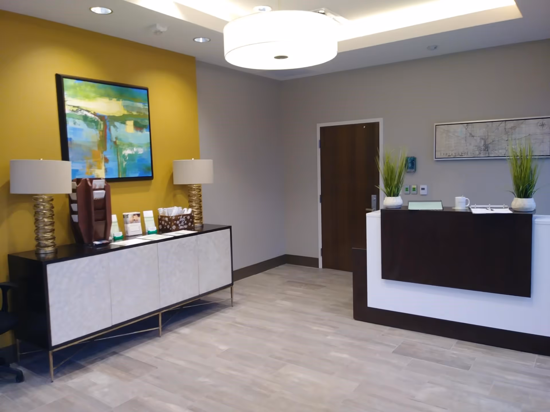 A modern reception/lobby area with a front desk, a sideboard topped with two lamps and artwork, and potted plants.