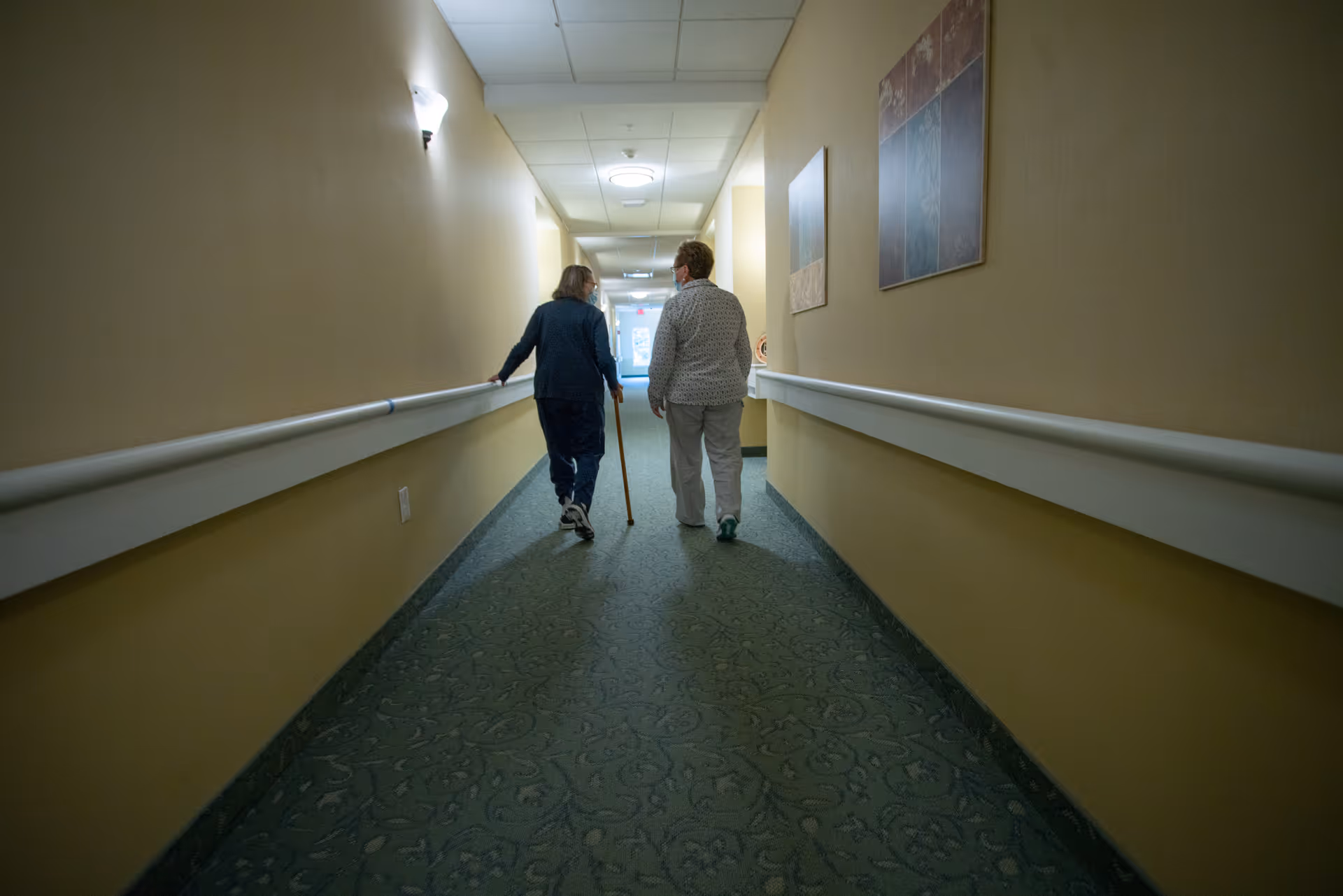 Two elderly women walking down a carpeted hallway with handrails on both sides in an assisted living facility. One woman is using a cane for support. The walls are beige with framed artwork, and the hallway is lit with ceiling lights.