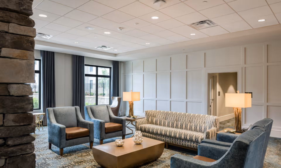 Well-lit communal lounge with a patterned sofa, blue armchairs, a coffee table, lamps, and large windows.