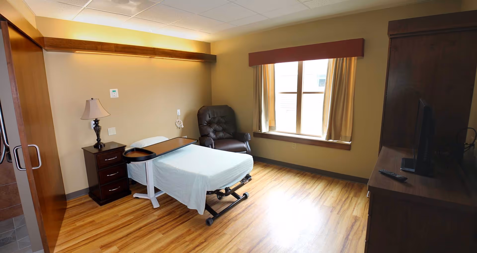 A simple, clean bedroom in a senior living facility with a single bed covered in white sheets, a bedside table with a lamp, a brown recliner chair near a window with beige curtains, and a wooden dresser with a TV on top. The room has wooden flooring and soft yellow walls.