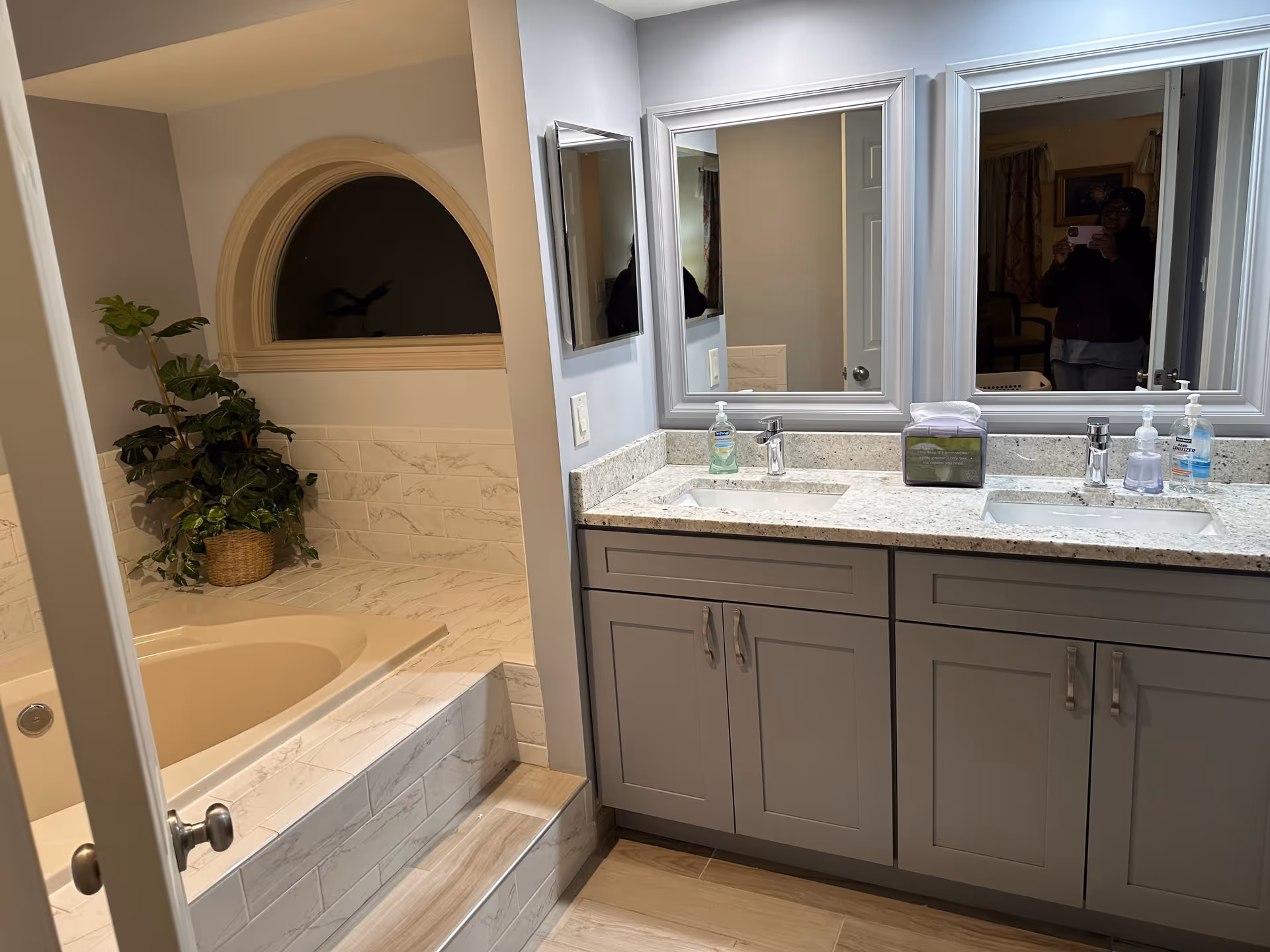 A bathroom with a double sink vanity featuring granite countertops and gray cabinets. Above the sinks are two large mirrors. To the left, there is a built-in bathtub with marble-like tile surround and a decorative arched window above it. A potted plant is placed on the bathtub ledge. The floor is covered with light wood-like tiles.