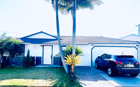 A single-story white building with a driveway, palm trees, and a parked dark SUV.
