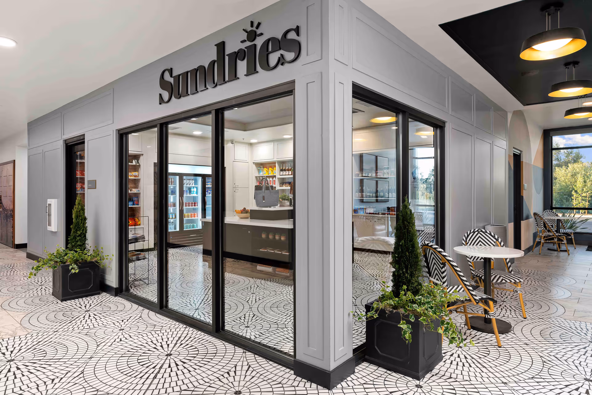 Interior corner view of a sundries shop within a senior living facility. The shop has large glass windows and doors, with shelves stocked with various items and refrigerated drinks visible inside. Outside the shop, there are small tables with black and white patterned chairs and potted plants on a tiled floor with a circular mosaic pattern. The area is well-lit with modern ceiling lights and large windows showing greenery outside.