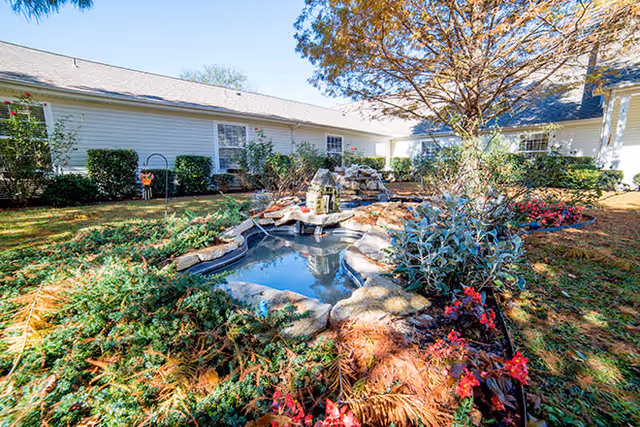 Sunlit courtyard with a small decorative pond and landscaped plants in front of a single-story building.