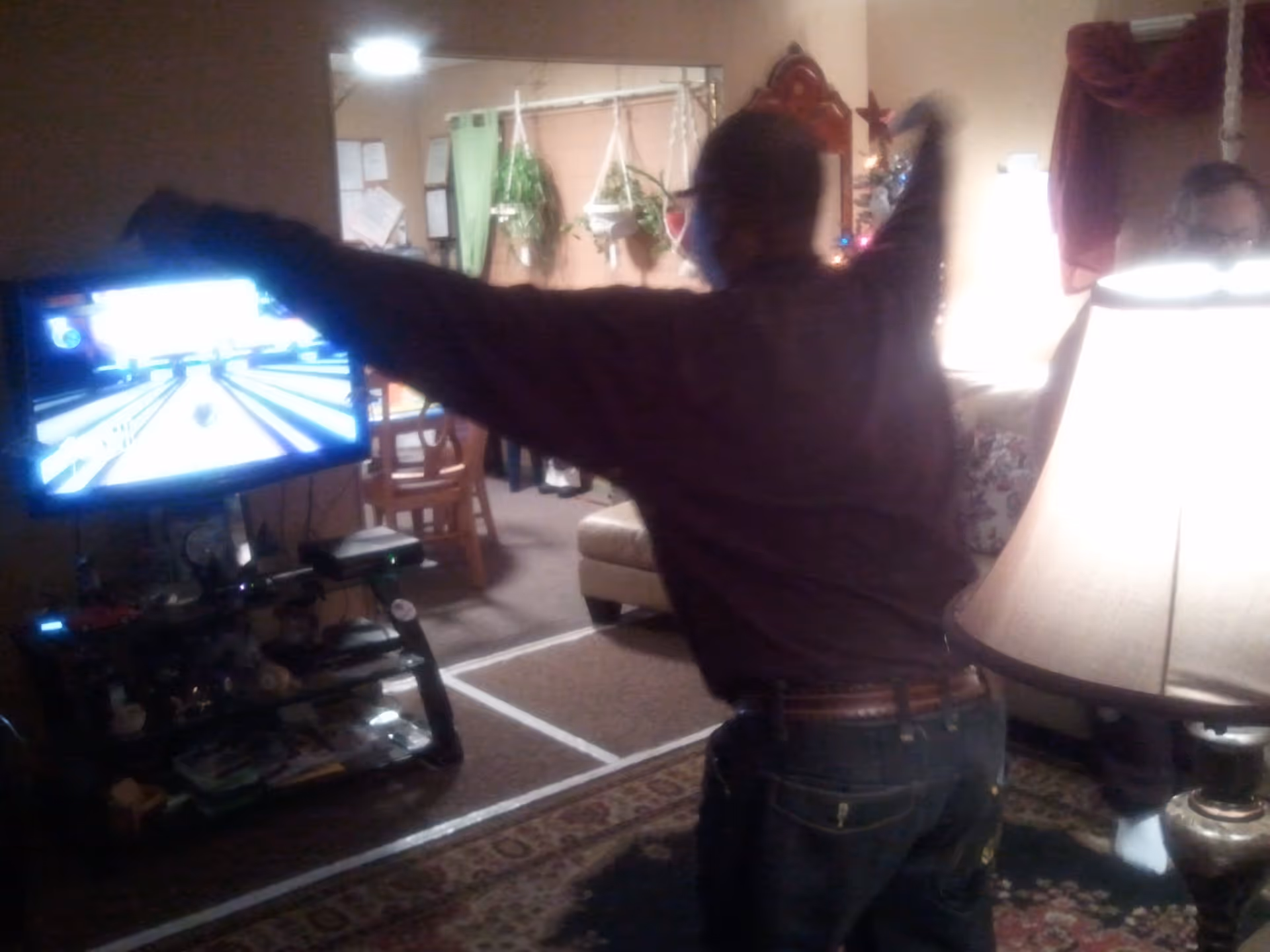 A person is playing a bowling video game on a television in a cozy living room. The person is in mid-motion, appearing to bowl, with arms extended. The room has a patterned rug, a lamp, a couch, and some hanging plants in the background.