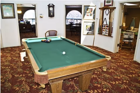 A green-felt pool table sits in a carpeted common room with a wall clock, cue rack, and doorways to adjoining rooms.