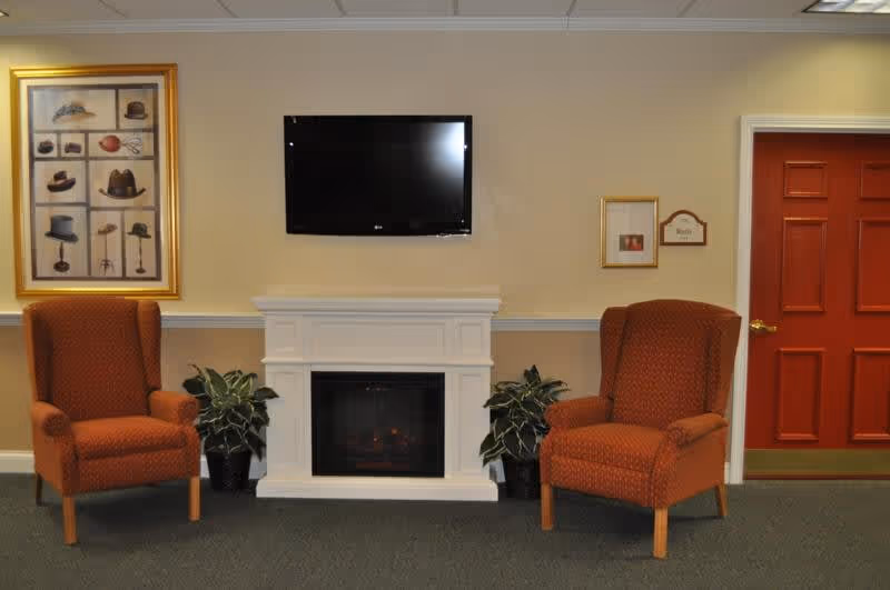 A cozy interior room with two orange upholstered armchairs placed on either side of a white electric fireplace. Above the fireplace is a flat-screen TV mounted on the beige wall. There are two potted plants flanking the fireplace. On the left wall, there is a framed picture of various hats, and on the right side, there is a red door and two small framed items on the wall.