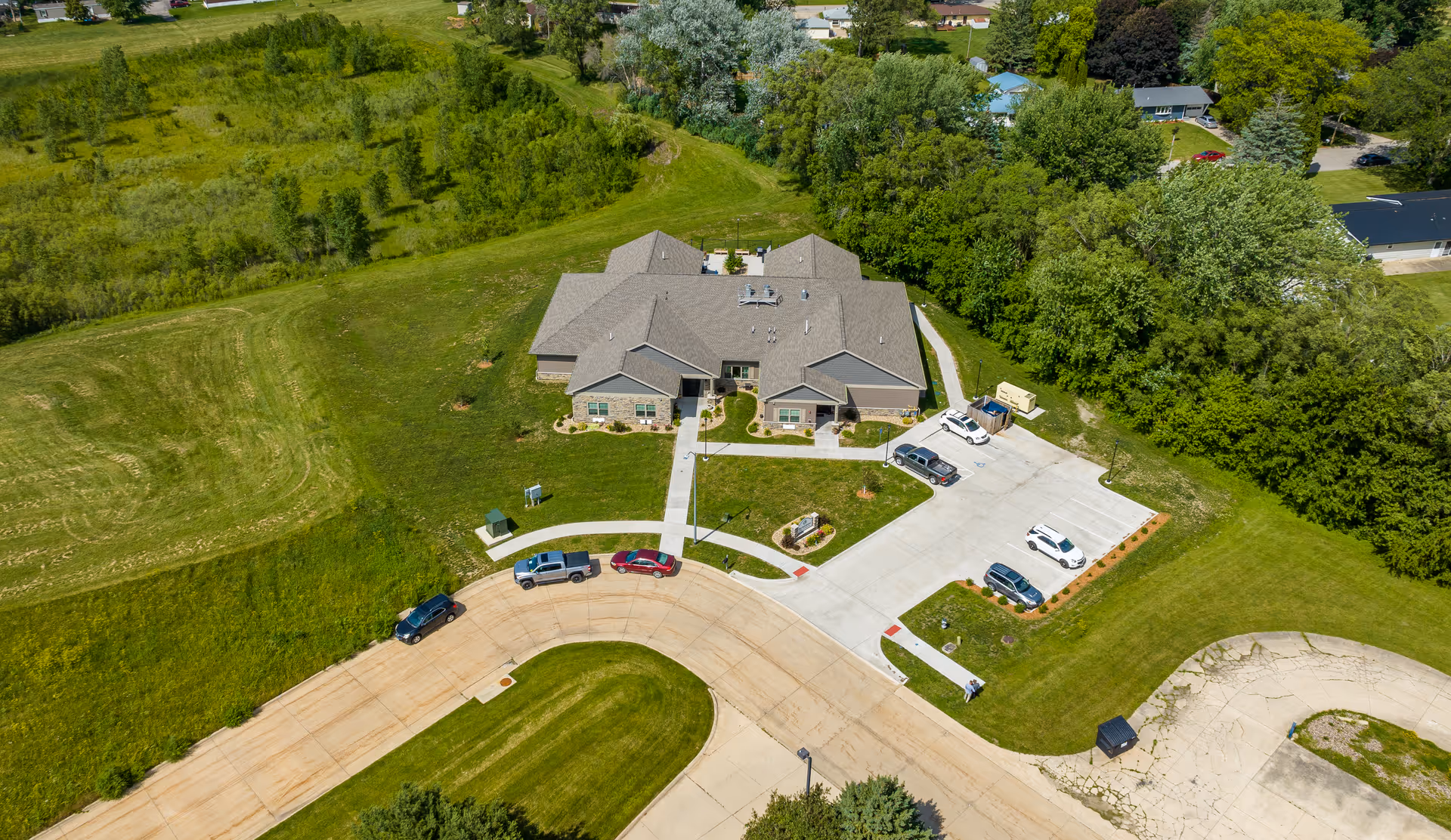 Aerial view of a single-story assisted living building with a circular driveway, parked cars, landscaped lawns and surrounding trees.