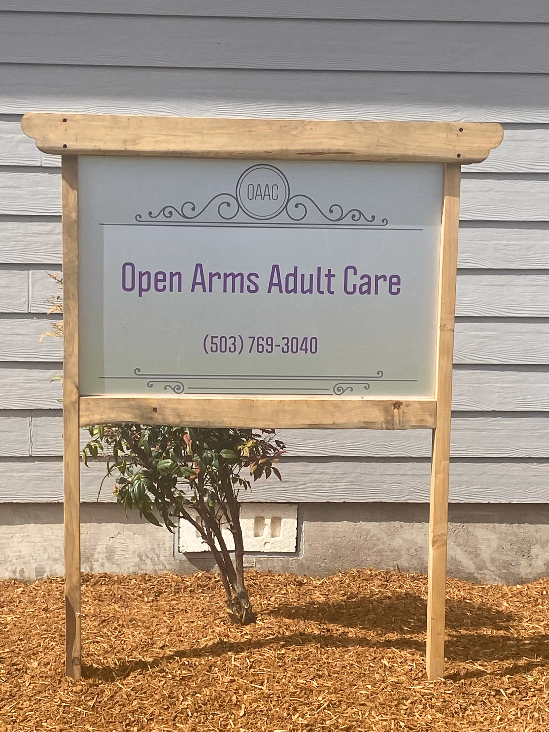 A wooden-framed sign in front of a gray building with horizontal siding. The sign reads 'Open Arms Adult Care' with the phone number (503) 769-3040 and a decorative logo with the letters OAAC at the top. There is a small bush and mulch at the base of the sign.