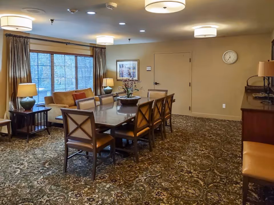 A cozy dining room with a large rectangular wooden table surrounded by eight cushioned chairs. The room features patterned carpet flooring, beige walls, three ceiling lights, two table lamps on side tables, a sofa near a large window with curtains, a wall clock, and framed artwork on the wall.