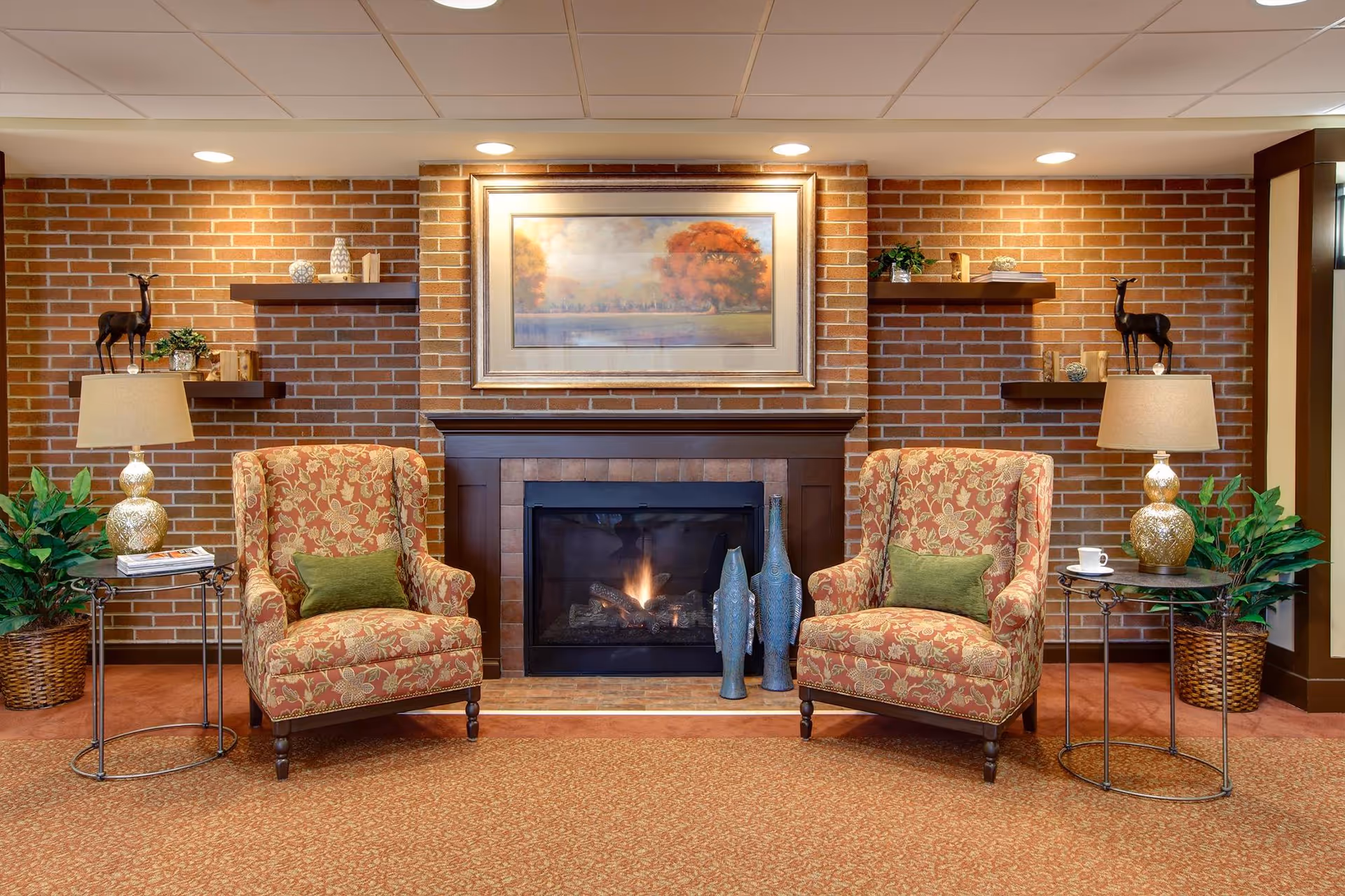 A cozy sitting area in a senior living facility featuring two patterned armchairs with green cushions placed on either side of a lit fireplace. The fireplace is set against a brick wall with a framed painting of an autumn tree above it. There are two side tables with lamps and decorative items, as well as potted plants on both sides. Floating shelves with small decorative objects are mounted on the brick wall.